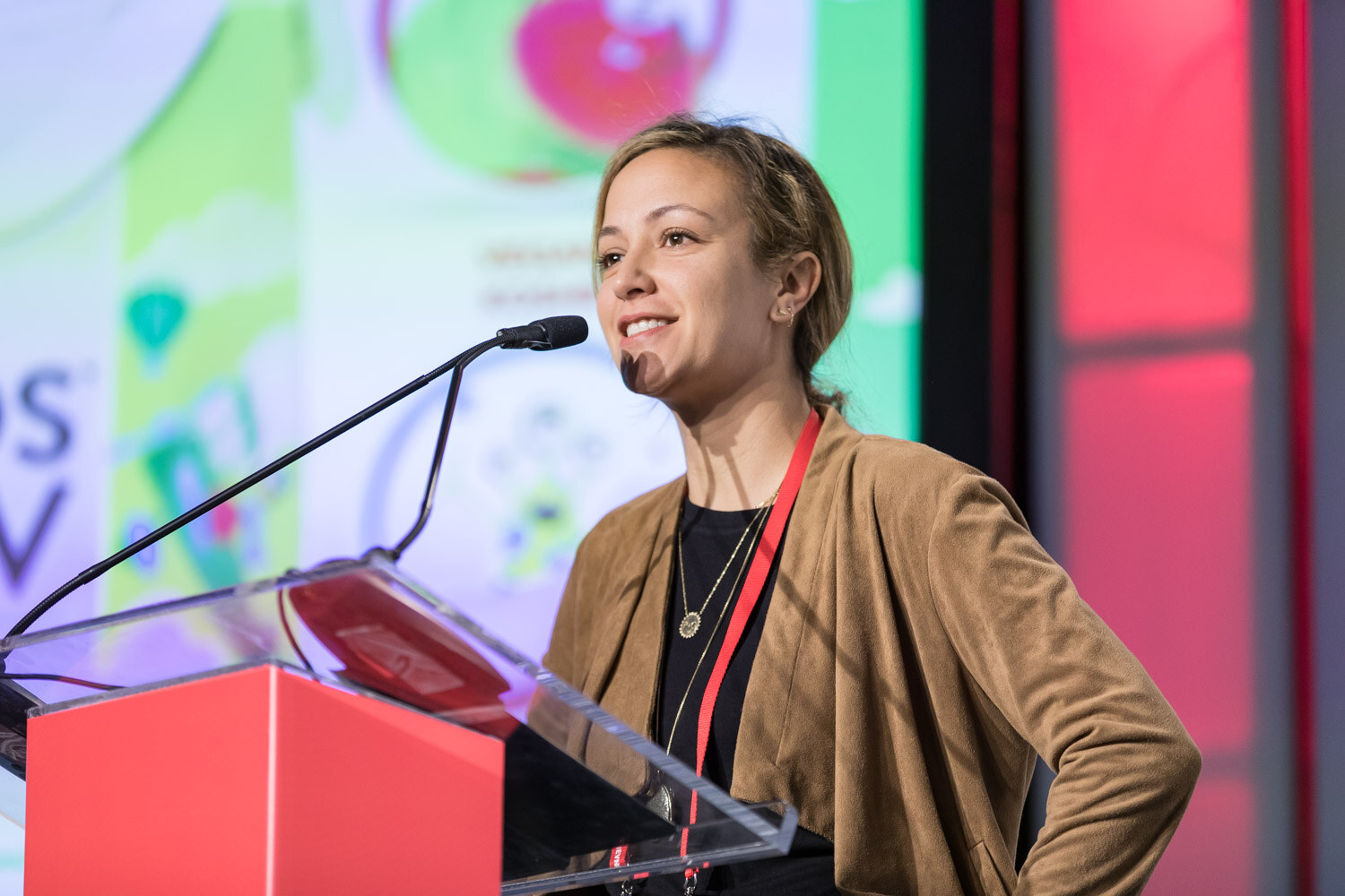 Woman in a tan jacket smiling at a podium with a Kids Luv slide in the background.