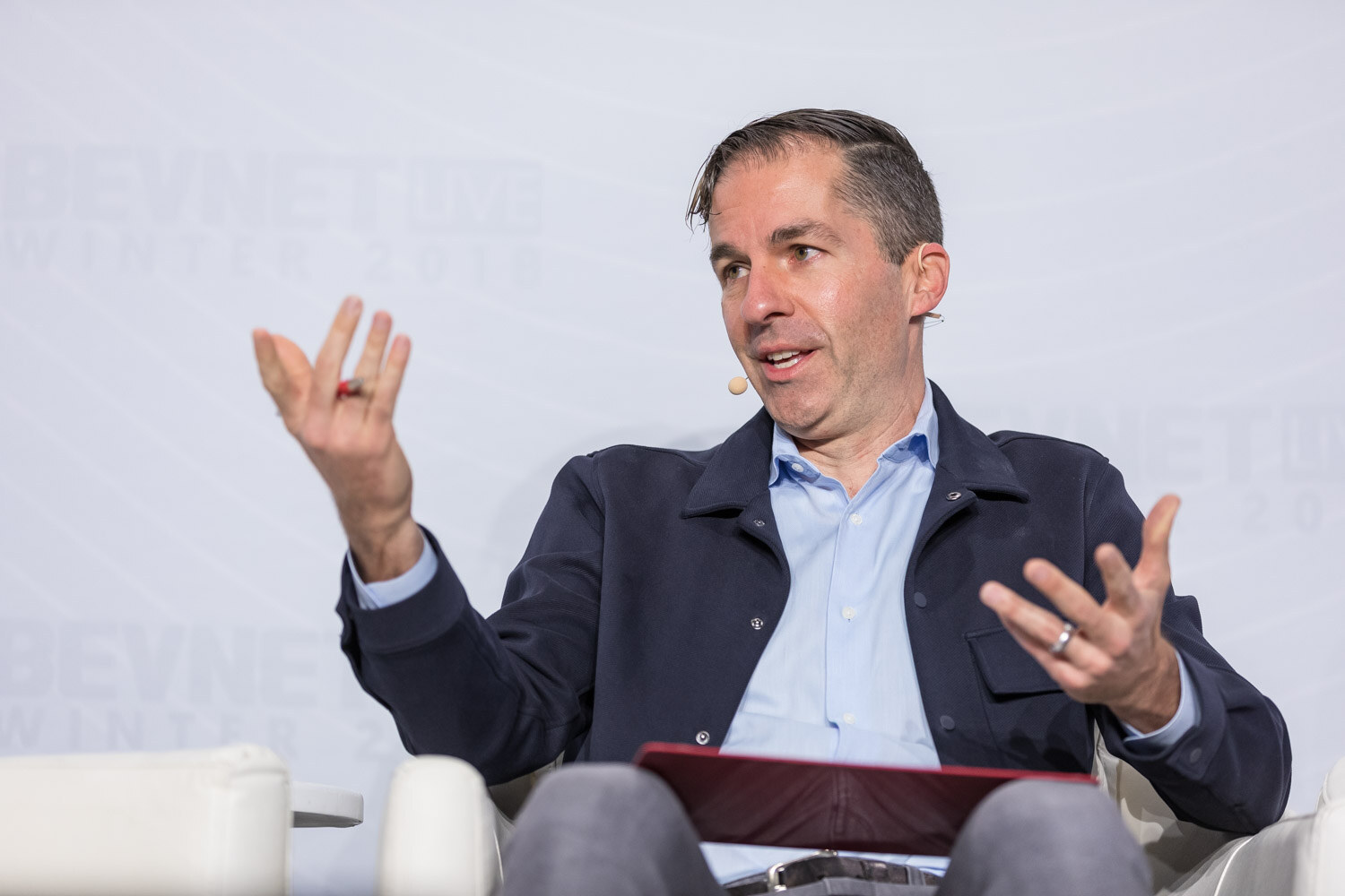 Man in a navy jacket and blue shirt gesturing with both hands while seated.