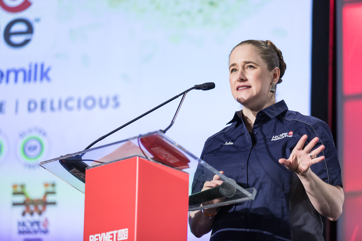 Woman in a navy work shirt speaking at a podium beside a Hope and Sesame slide.