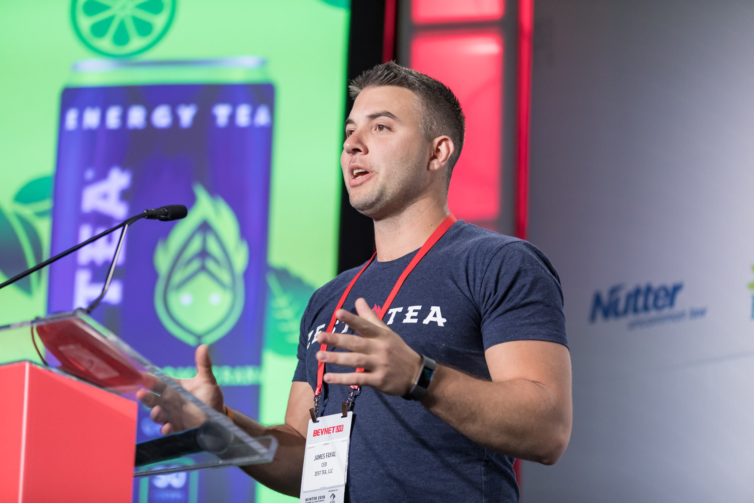 Man in a navy Zest Tea shirt speaking at a podium in front of a green slide.