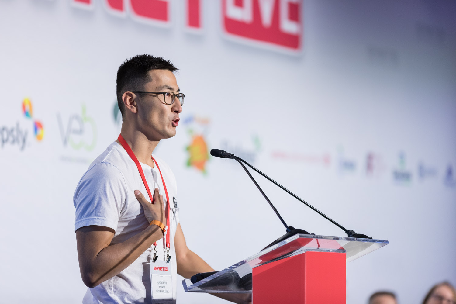 Man in glasses and a white t-shirt speaking at a clear podium on stage.