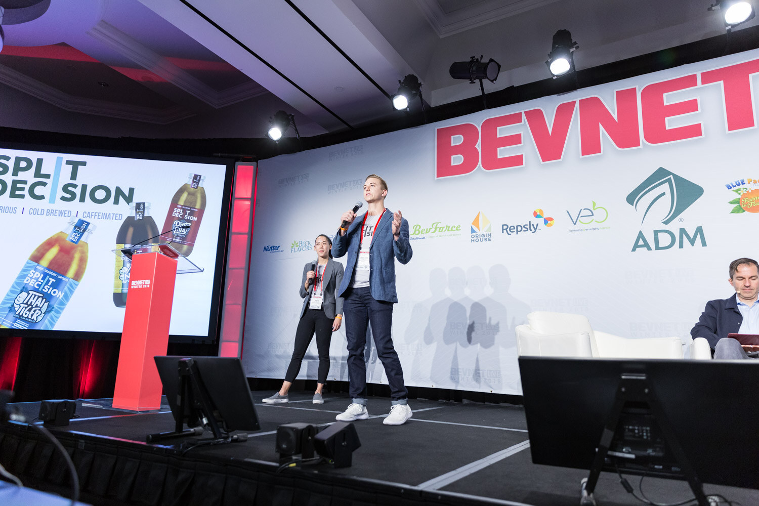 Man and woman standing on stage with a screen showing Split Decision bottles.