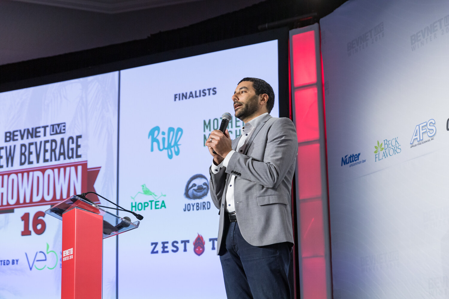 Man in a gray blazer holding a microphone beside a large screen reading New Beverage Showdown 16 Finalists.