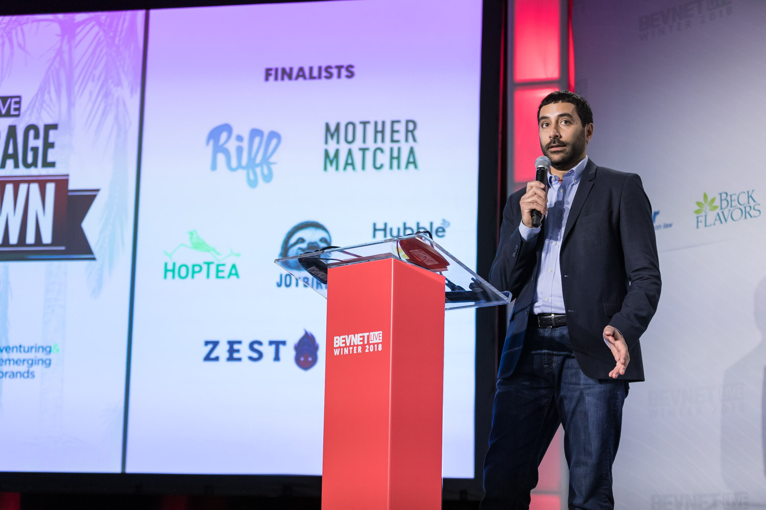Man in a dark blazer holding a microphone beside a screen showing a lineup of Showdown finalist logos.
