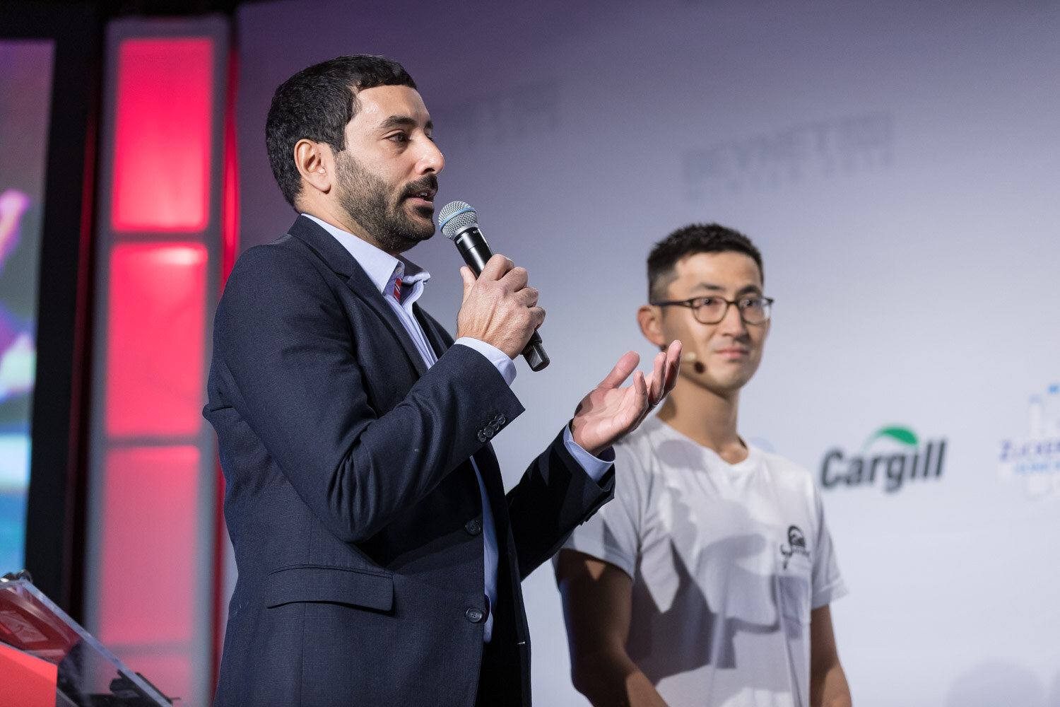 Man in a navy blazer holding a microphone beside a man in a white t-shirt on stage.