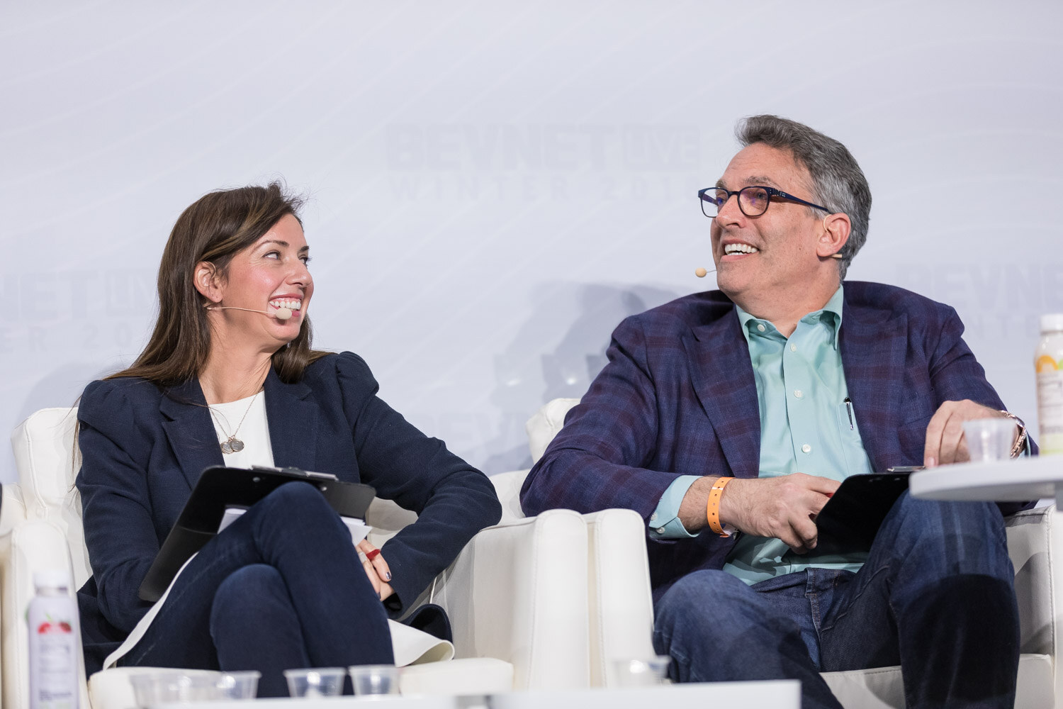 Woman in a navy blazer and a man in glasses seated on stage, both smiling.