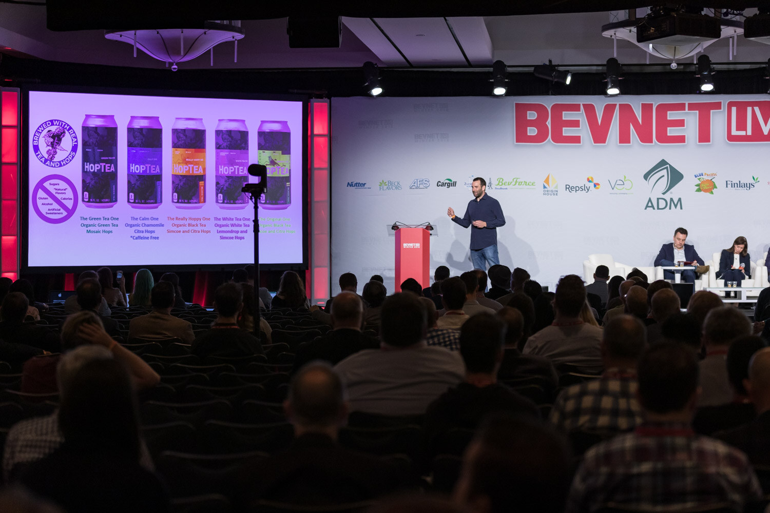 Large ballroom audience looking at a presenter on stage next to a screen of bottles.
