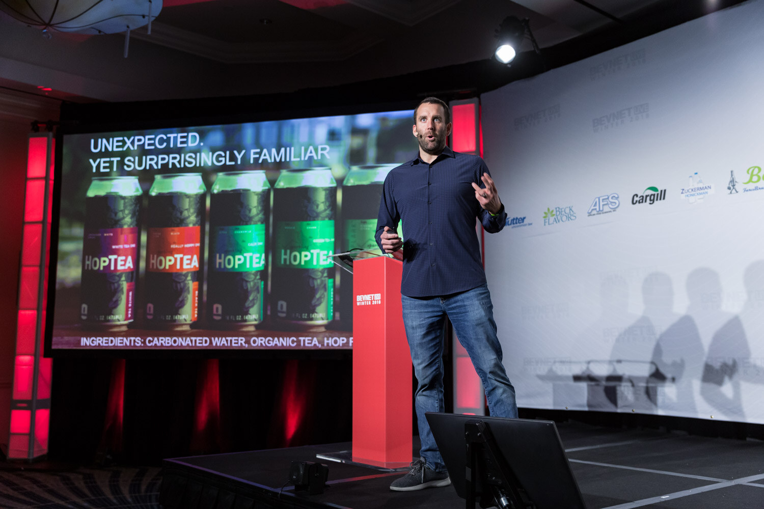 Man in a dark shirt on stage with a screen showing Hoptea cans and the word Unexpected.