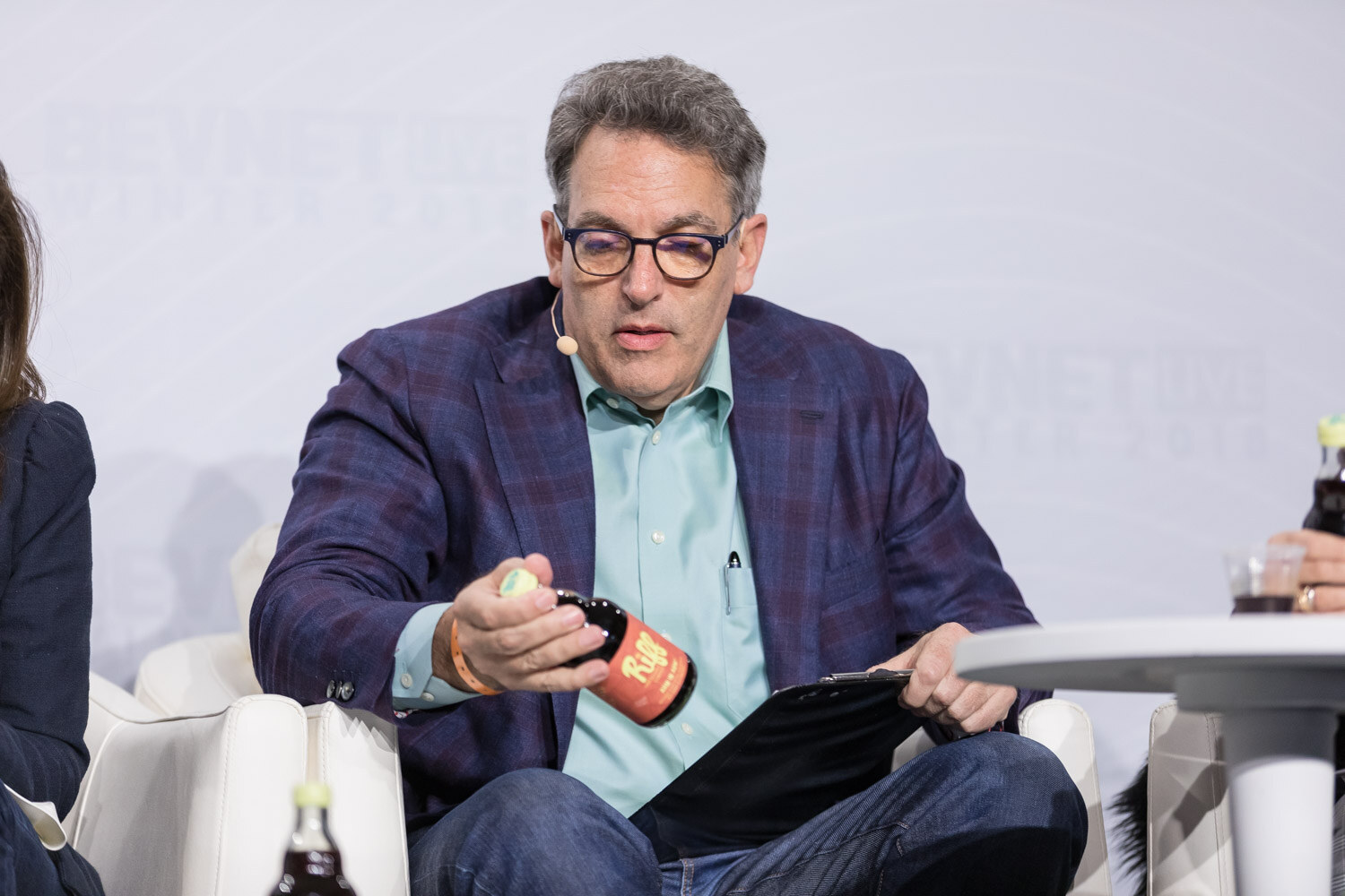 Man in a plaid blazer and glasses holding a small bottle on stage.
