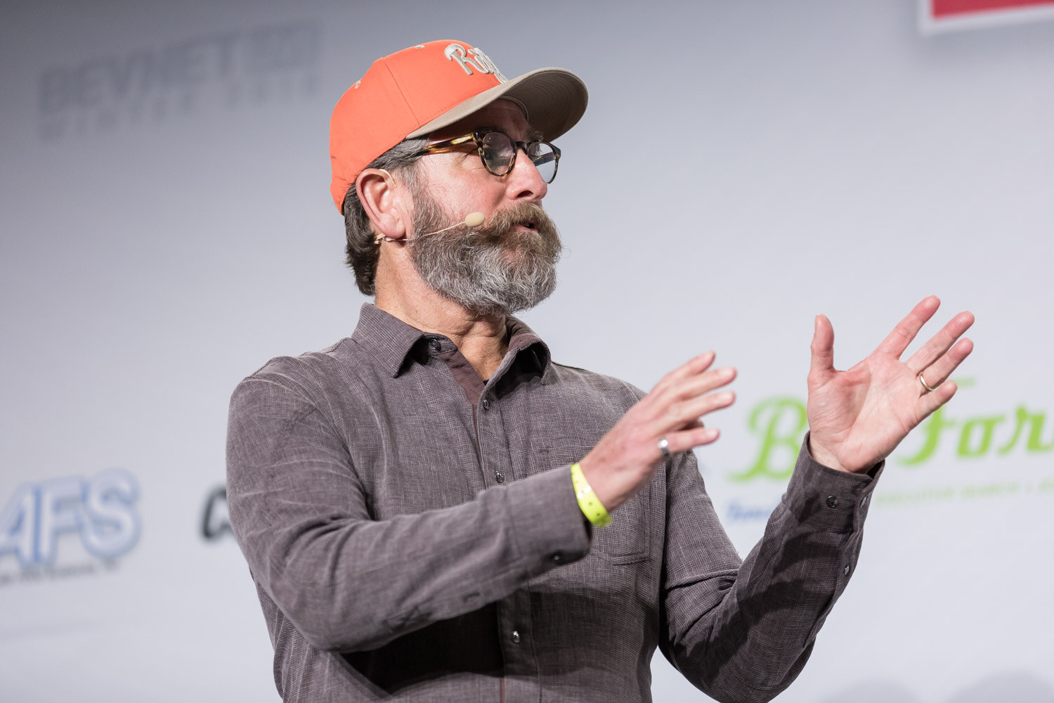 Bearded man in an orange cap and gray shirt gesturing on stage.