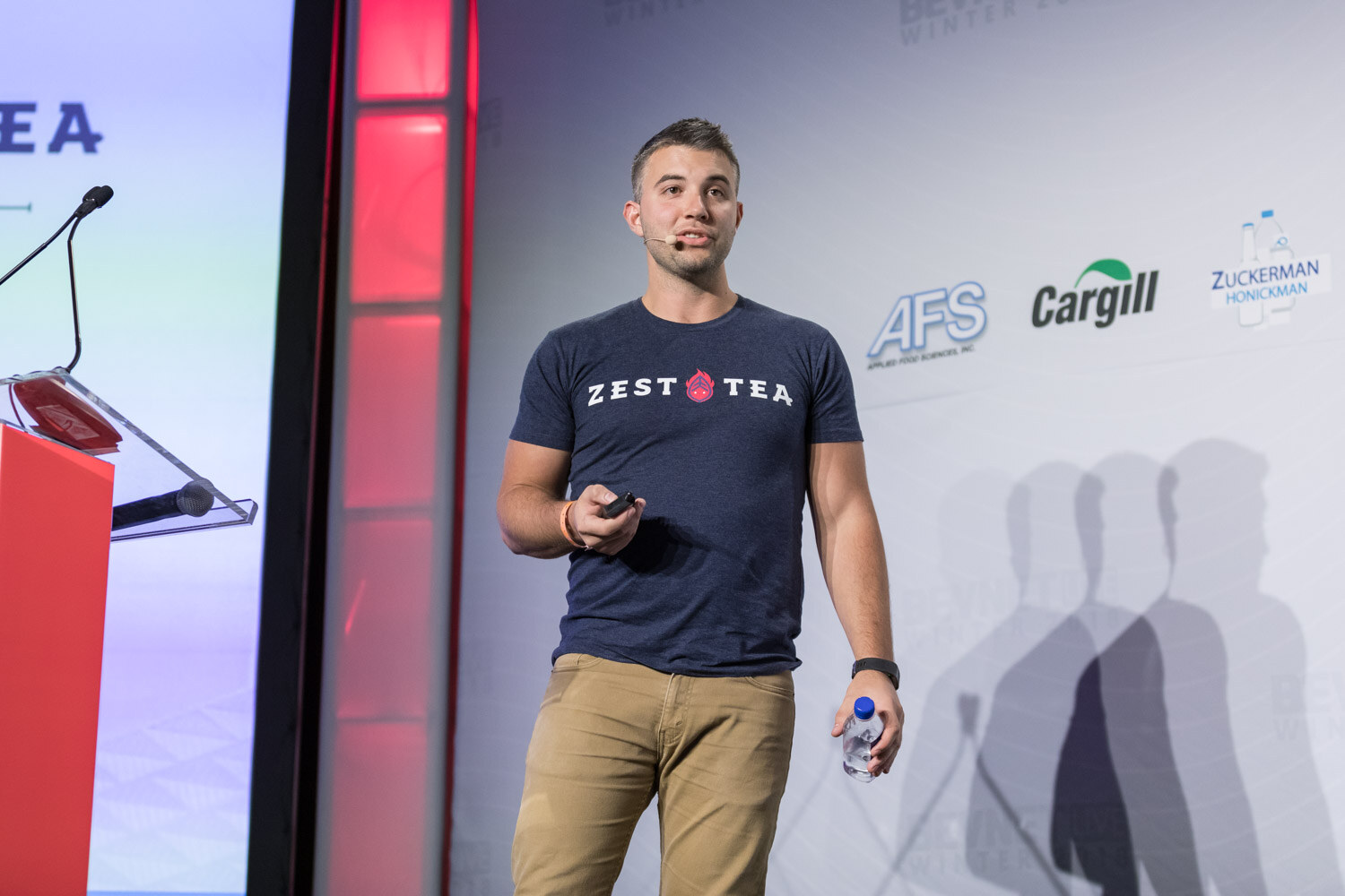 Man in a navy Zest Tea shirt and khakis standing on stage with sponsor logos behind him.
