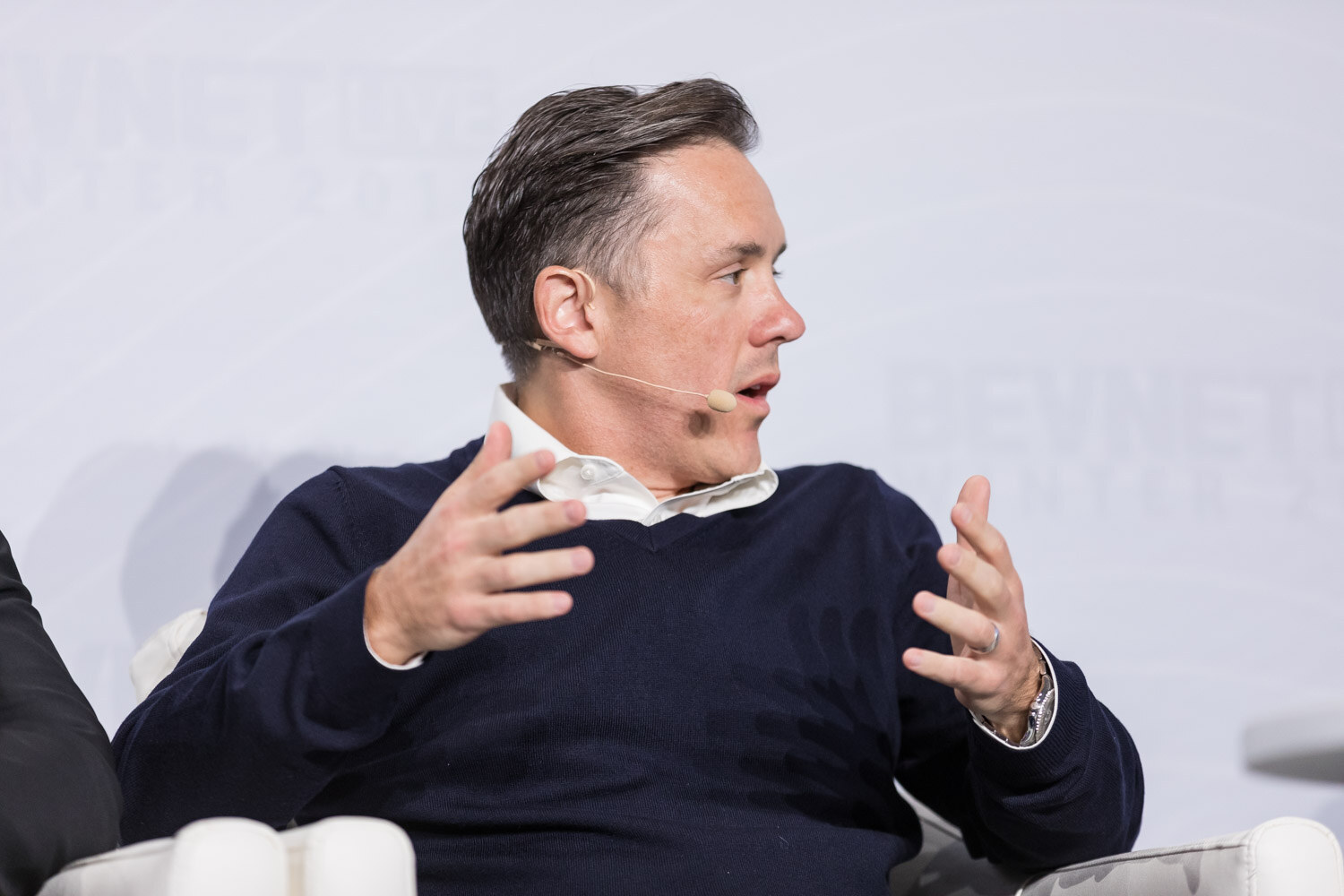 Man in a dark sweater and white collar gesturing while seated.