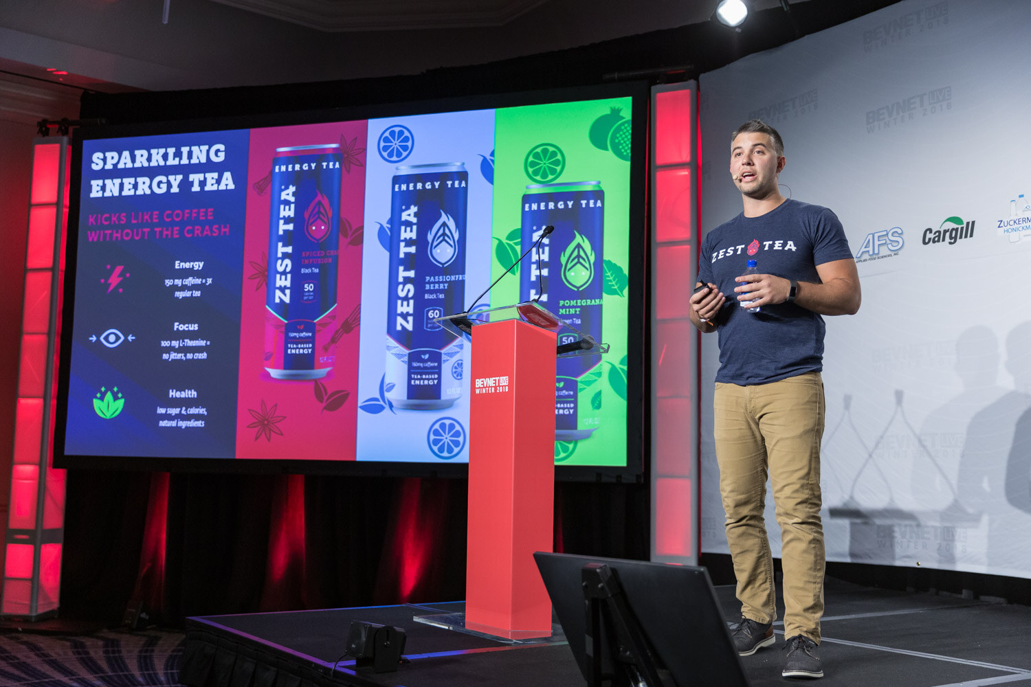 Man in a navy shirt on stage next to a screen showing Sparkling Energy Tea cans.