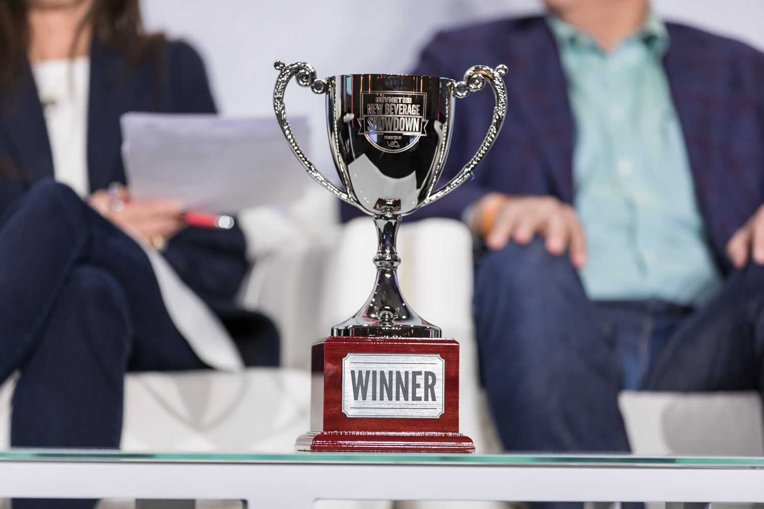 Silver trophy labeled Winner on a stage table with two seated people behind it.