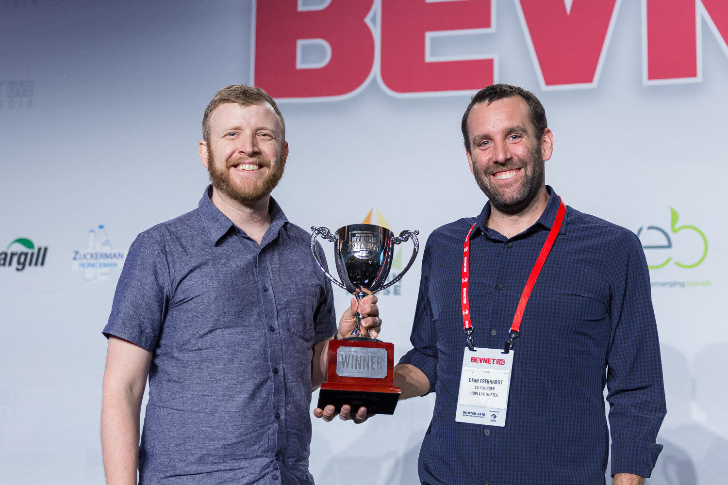 Two men with a silver trophy cup, one holding it in front of a BevNET Live backdrop.
