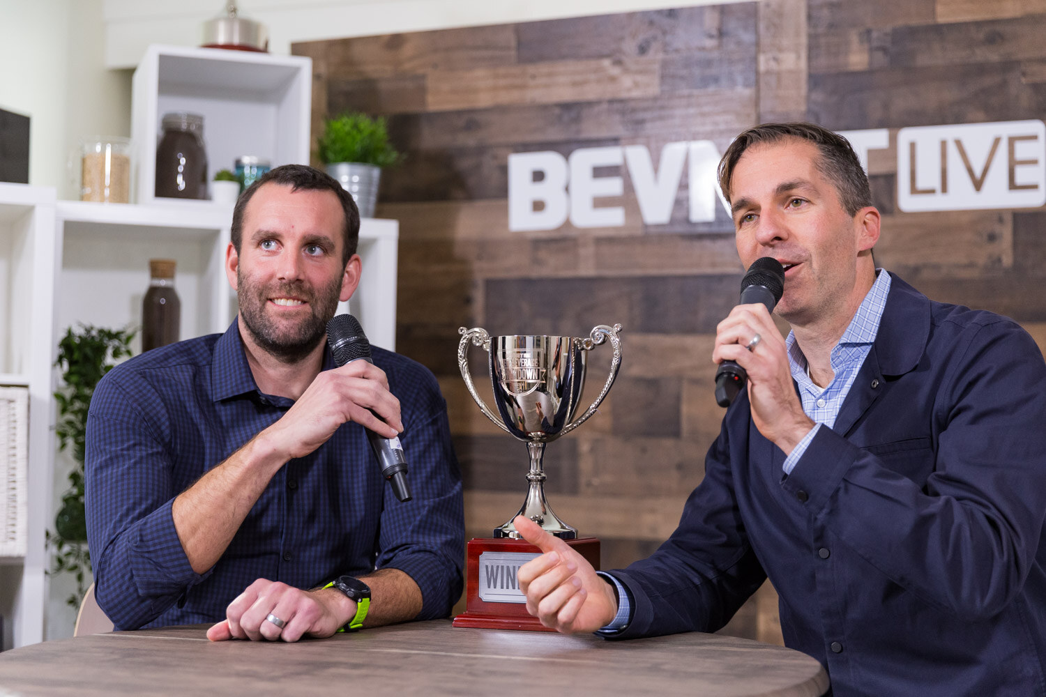 Two men holding microphones with a silver trophy on a table in front of a wood wall.