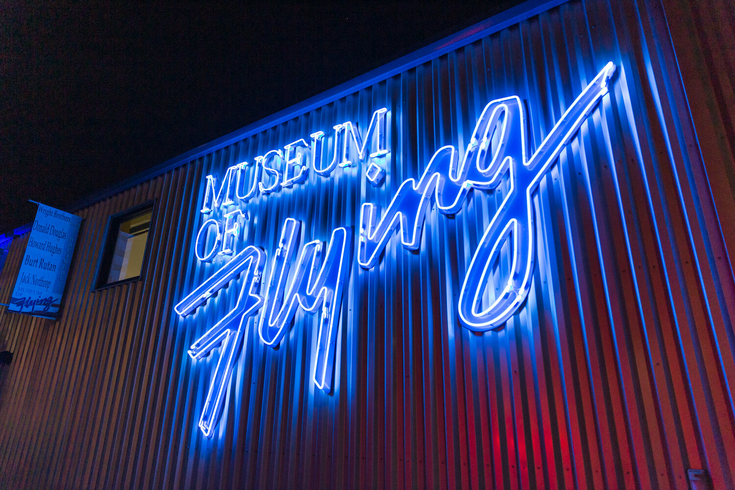 Blue neon sign reading Museum of Flying on a corrugated building at night.