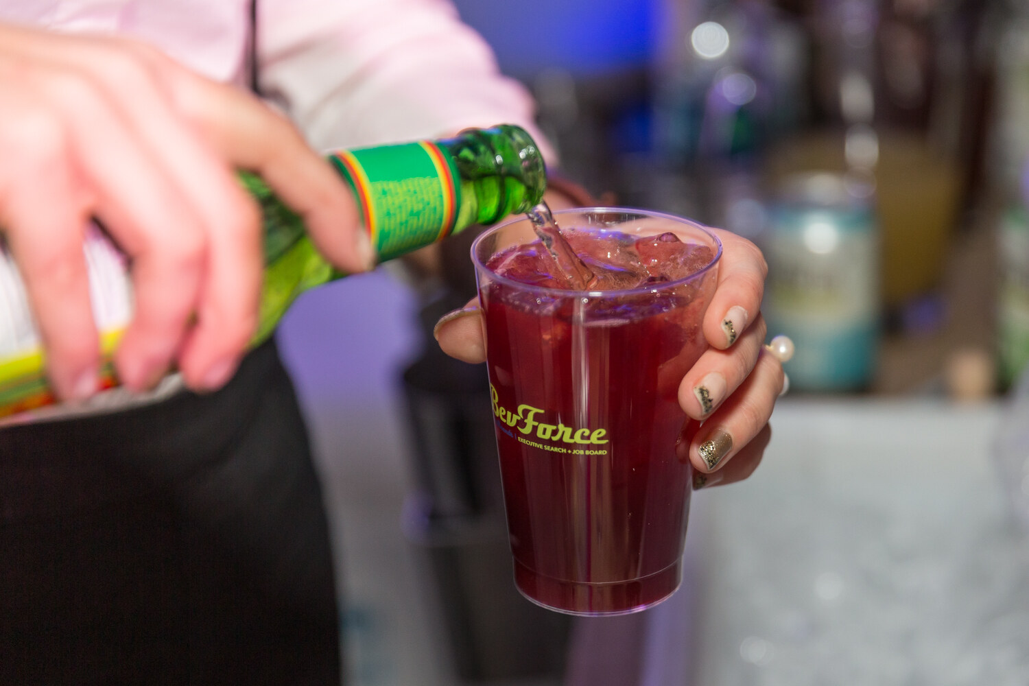 Hand pouring a green bottle into a clear cup of dark red liquid.