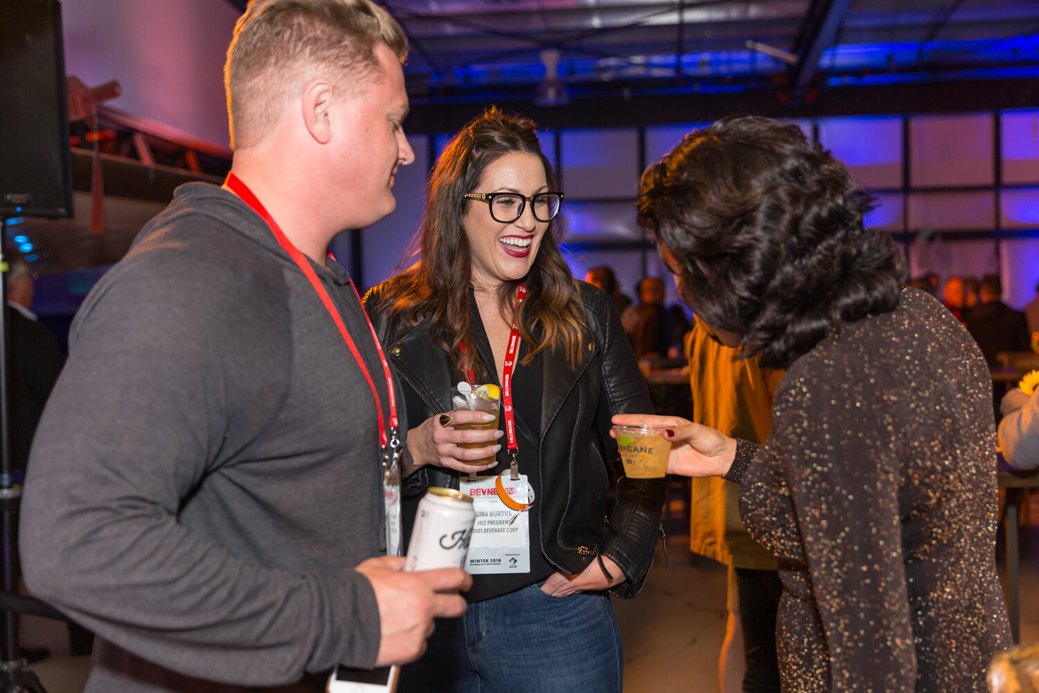 Man and two women with lanyards and cups talking in a dim hangar.