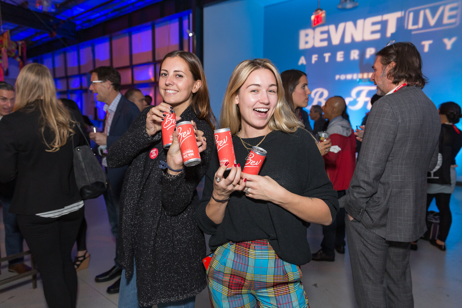 Two smiling women holding red cans while guests stand behind them.