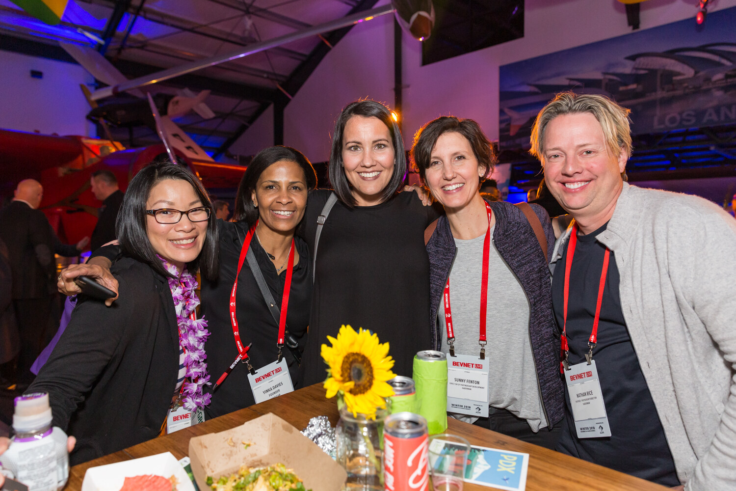 Five women with lanyards and leis smiling around a table with a sunflower centerpiece.