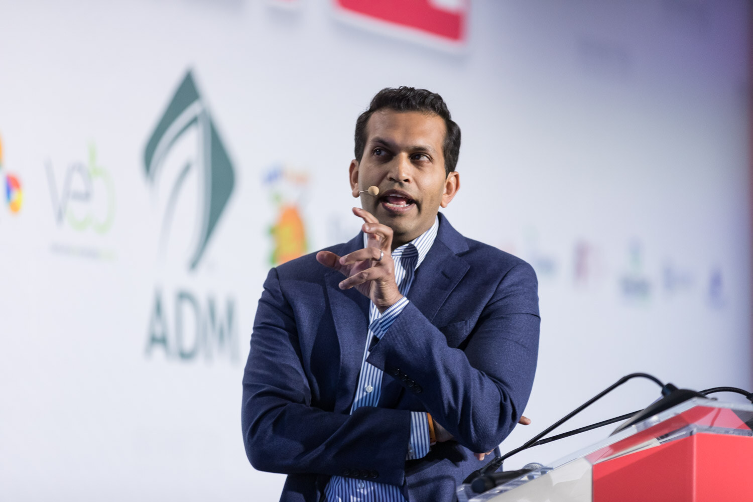 Man in a navy blazer gesturing in front of a white sponsor backdrop with an ADM logo.