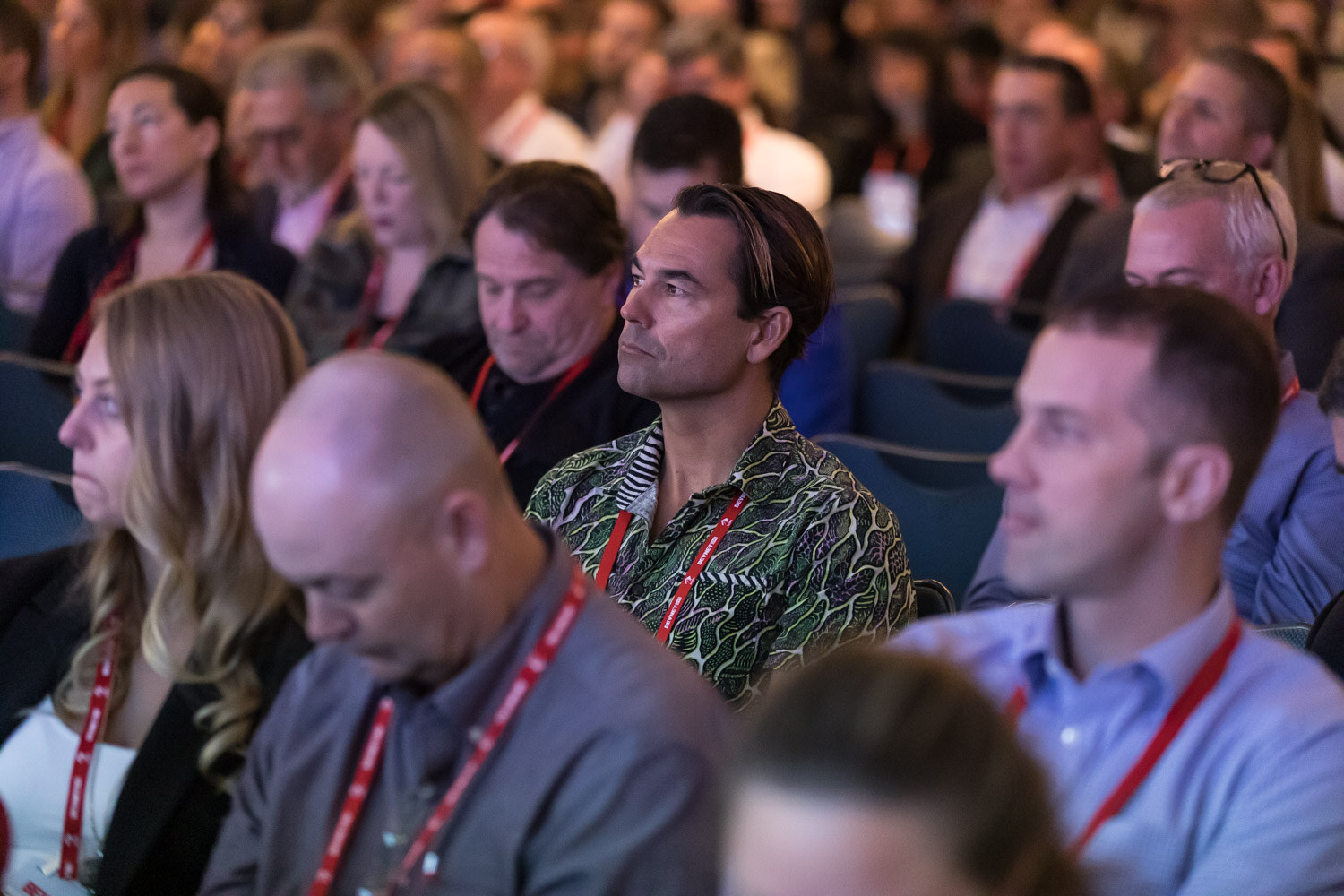 Close view of seated conference attendees with red lanyards watching the stage.