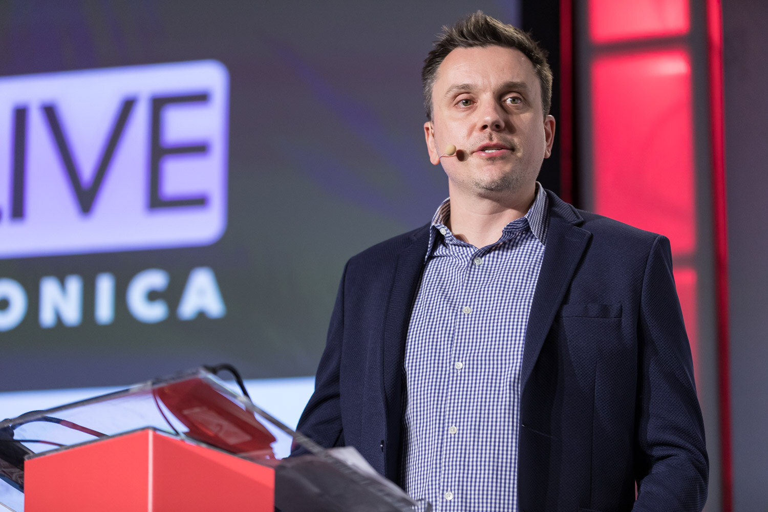 Man in a dark jacket speaking at a red podium in front of a Santa Monica event graphic.