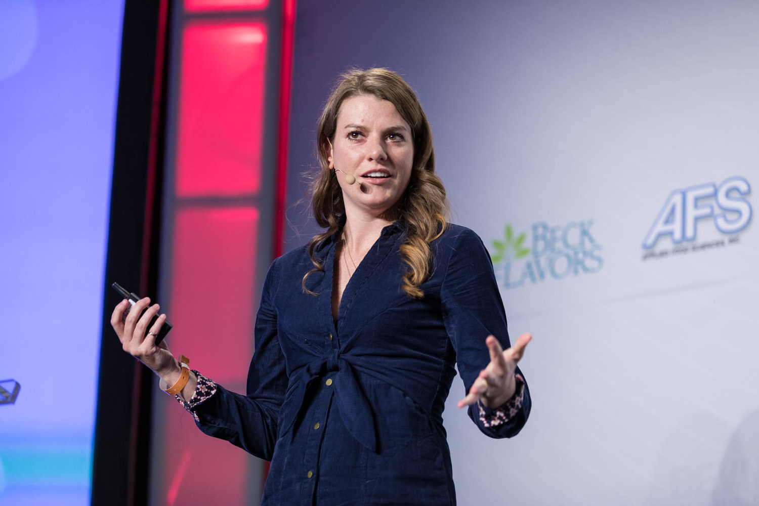 Woman in a navy dress presenting on stage with sponsor logos behind her.
