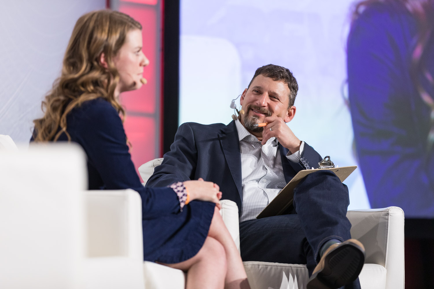 Woman and man talking in white armchairs on stage, the man laughing and holding a clipboard.