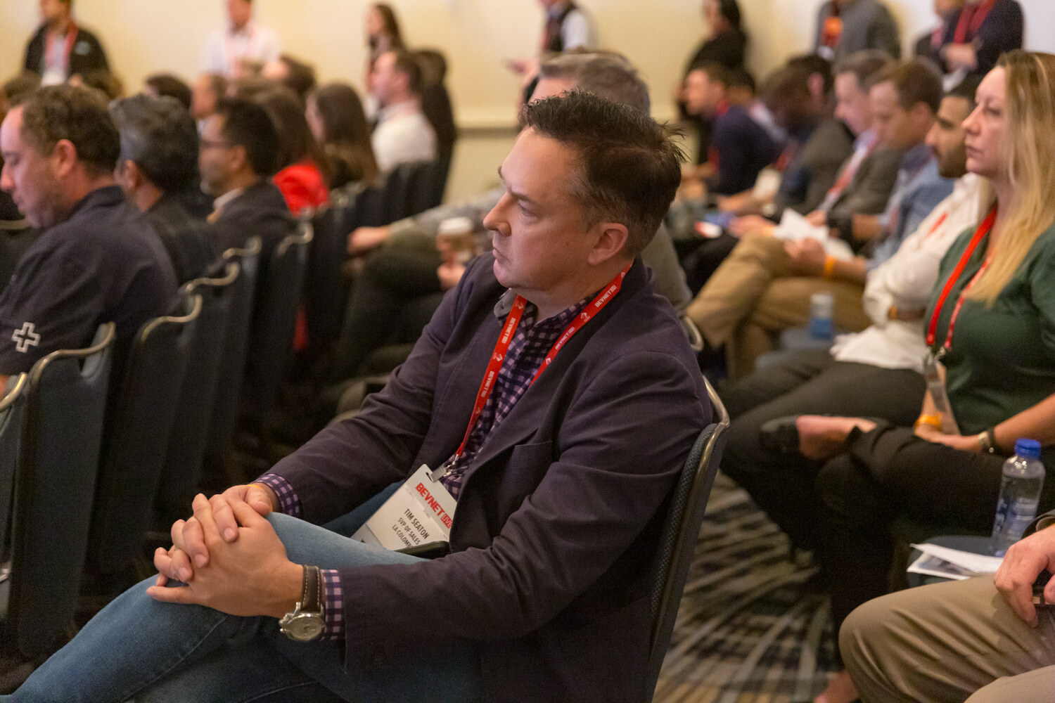 Man in a dark blazer with a lanyard seated in an audience looking toward the front of the room.