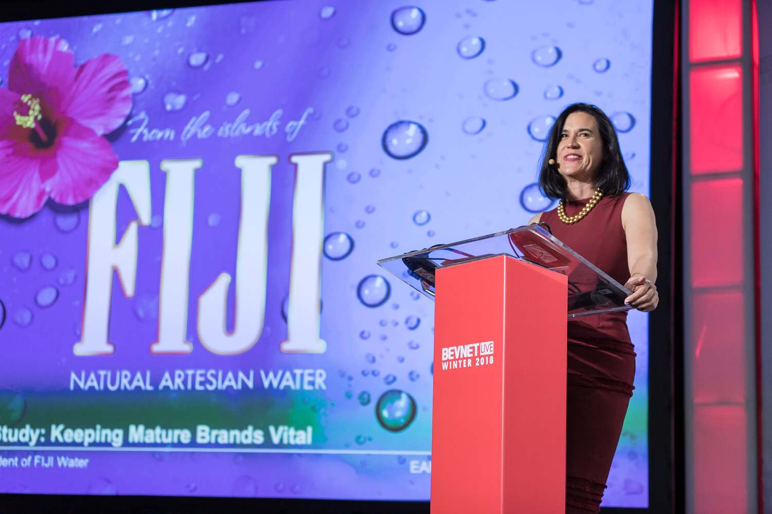 Woman in a red dress at a podium next to a screen showing the Fiji Natural Artesian Water logo.