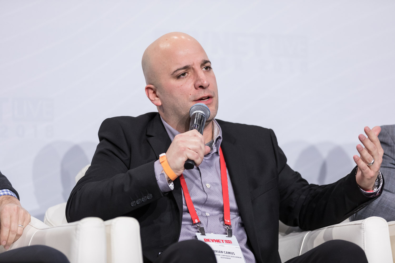 Bald man in a dark blazer speaking into a handheld microphone.