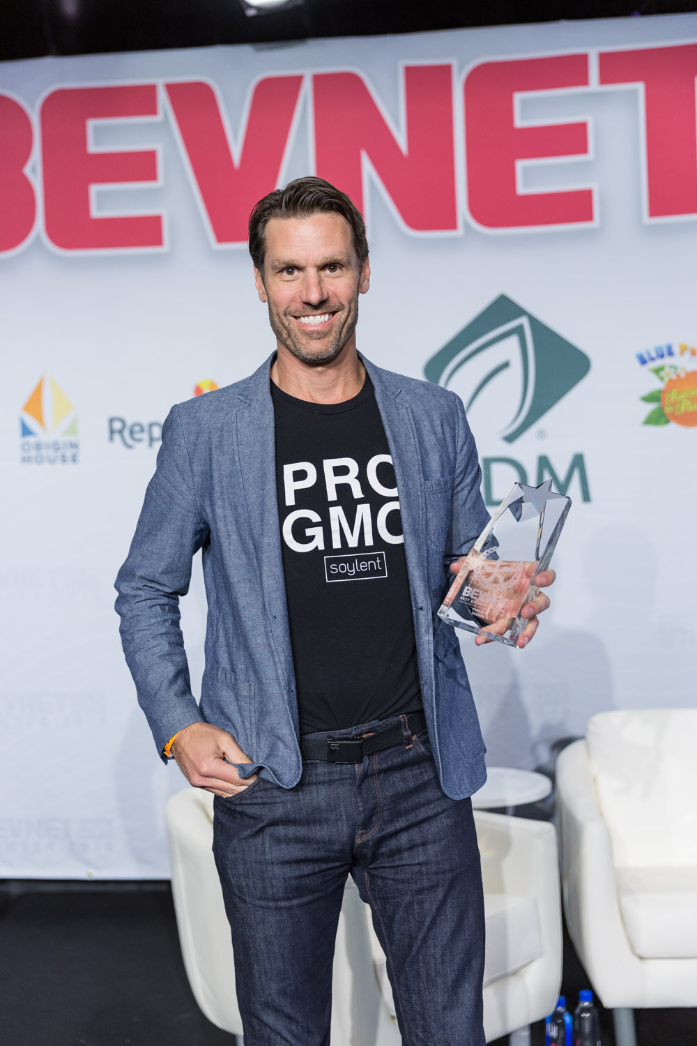 Man in a blazer and a graphic t-shirt holding a glass award on a stage with a BevNET Live backdrop.