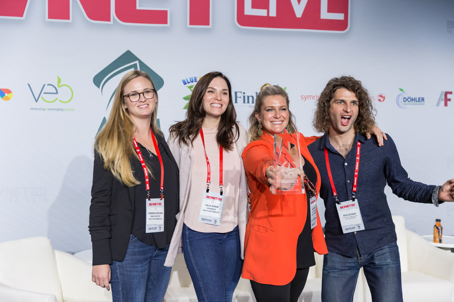 Four people with arms around each other on stage, one holding a glass award.