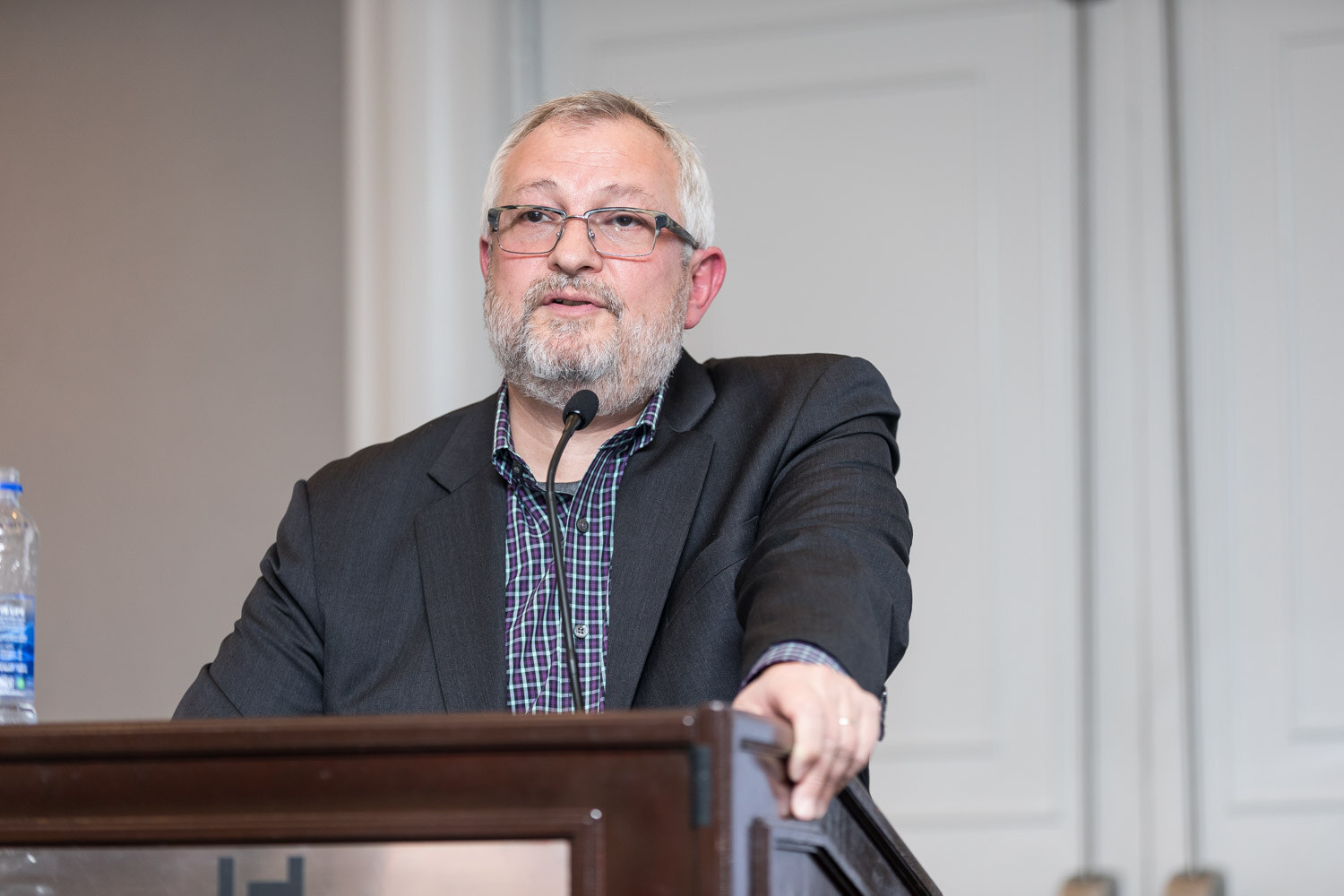 Gray-bearded man in glasses and a dark blazer speaking at a wooden lectern.