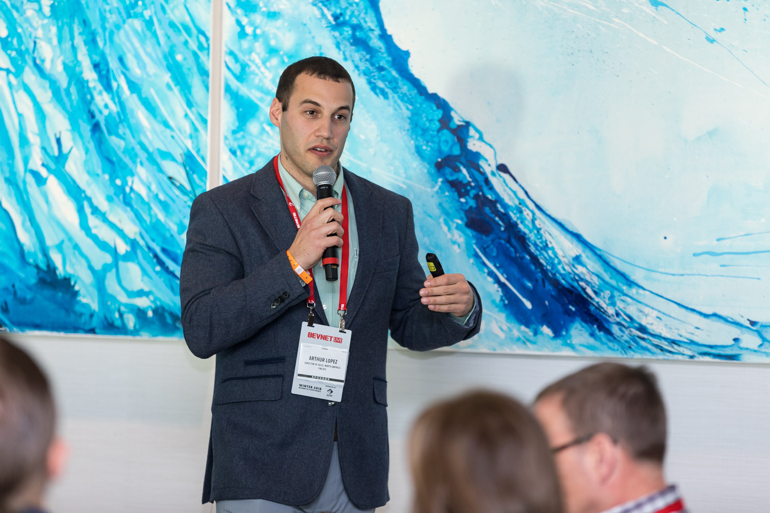 Man in a navy blazer with a microphone and a slide clicker in front of a blue wave painting.