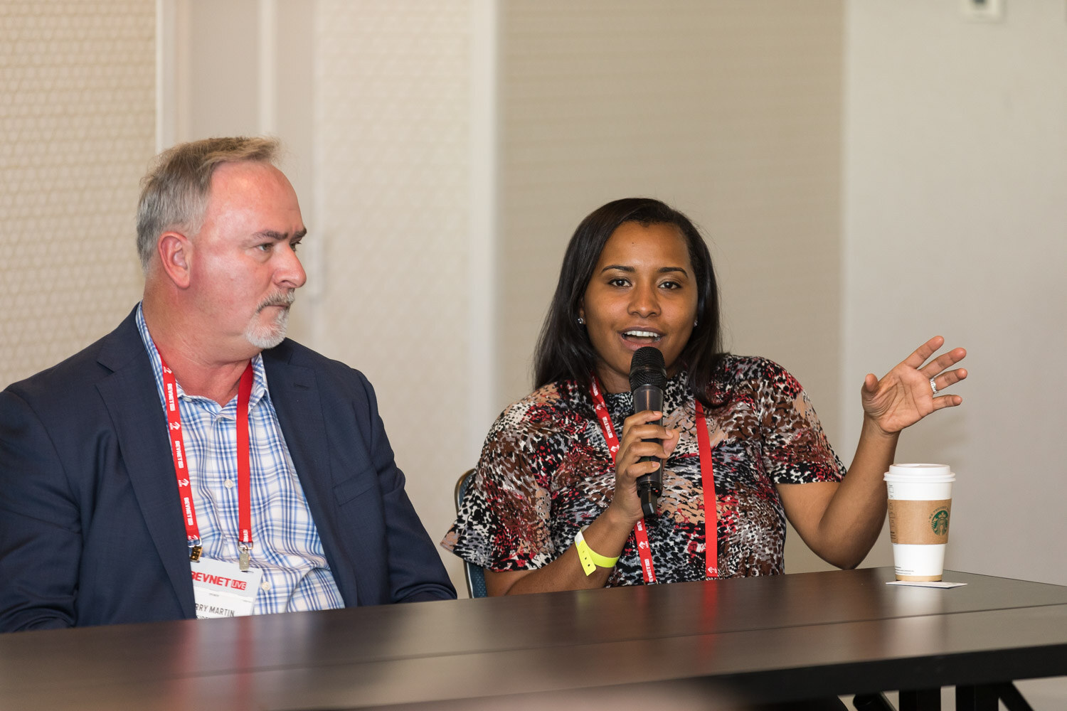 Three people at a long table, a woman in a patterned top holding a microphone and gesturing.