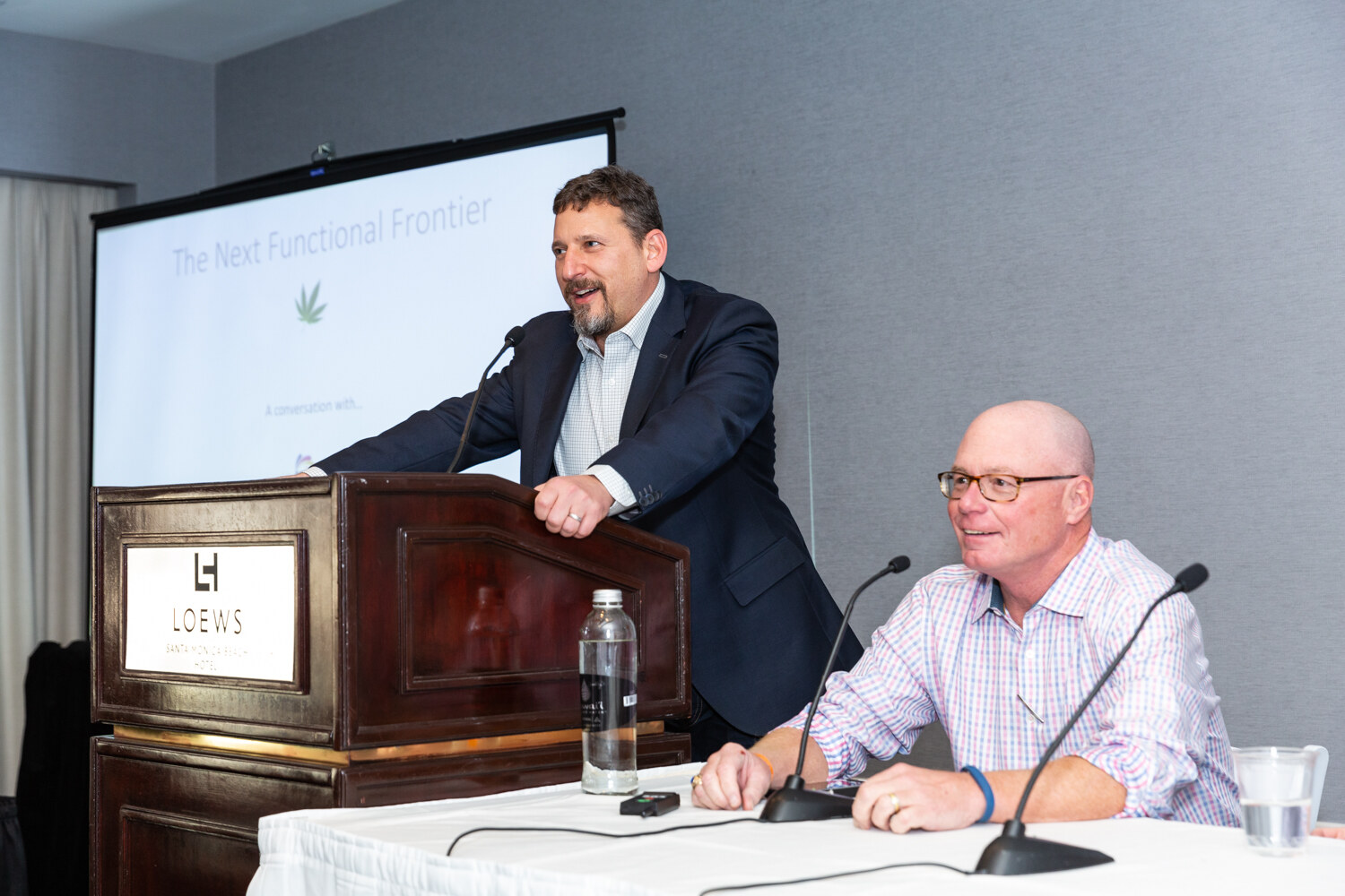 Man at a lectern and a second man seated at a table in front of a projector screen.