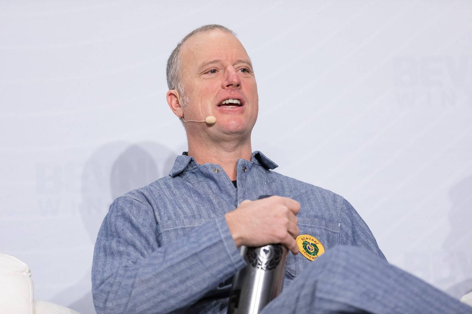 Man in a chambray shirt holding a metal water bottle in an armchair on stage.