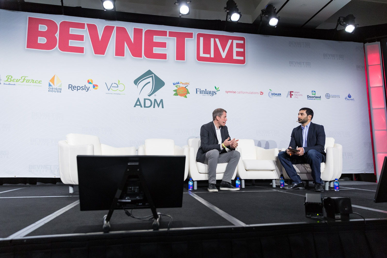 Two men seated in white armchairs on a stage in front of a BevNET Live backdrop.