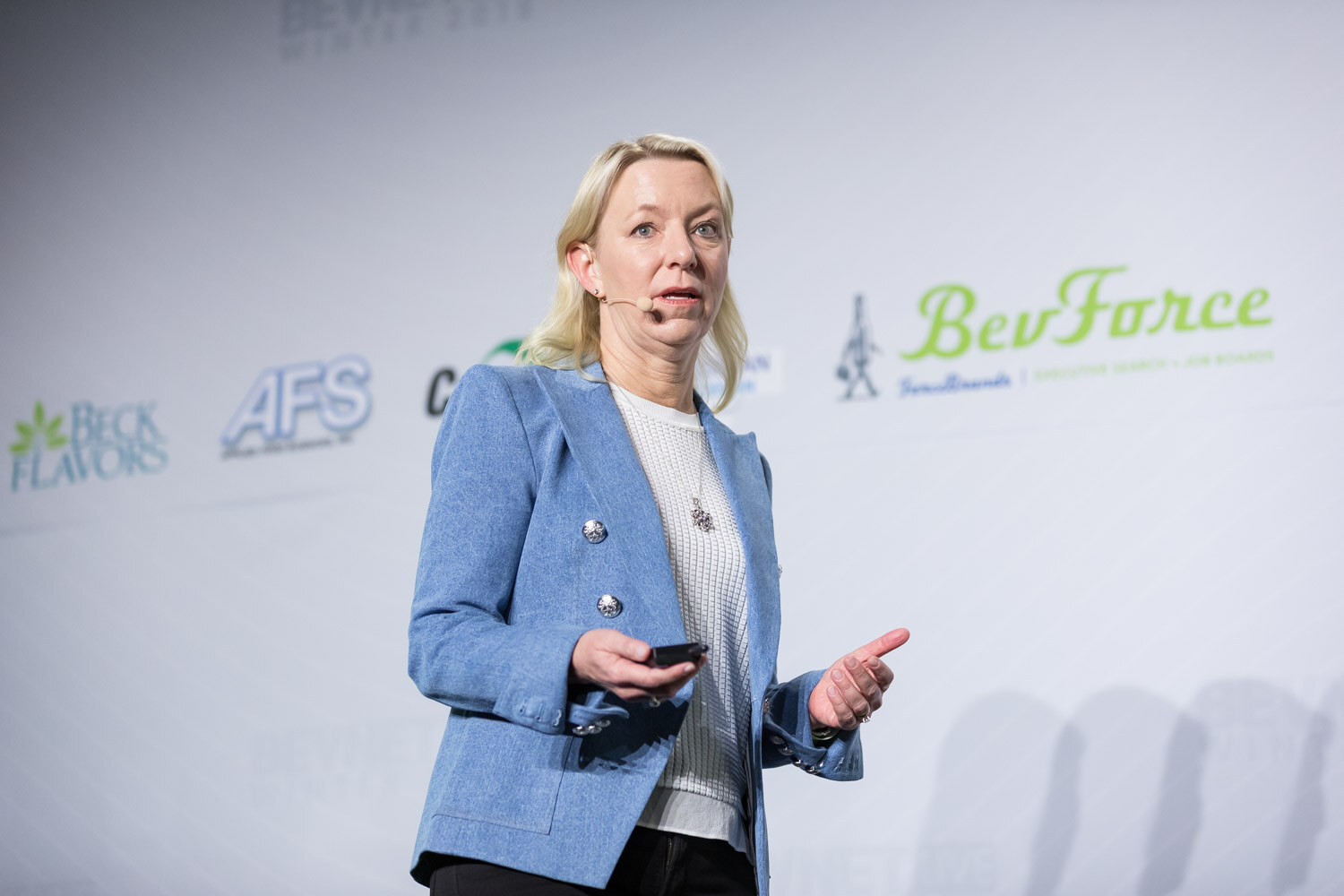 Woman in a light blue blazer holding a clicker on stage with sponsor logos behind her.