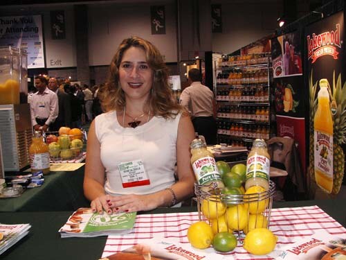 A smiling woman in a white sleeveless top stands behind a table holding Lakewood juice bottles, a basket of limes and lemons, and brochures, with juice shelves behind her.