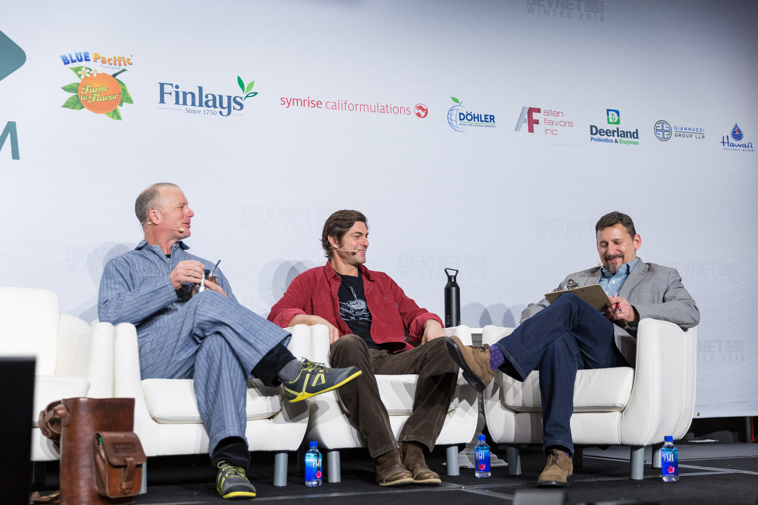 Three men seated on white lounge furniture on a stage.