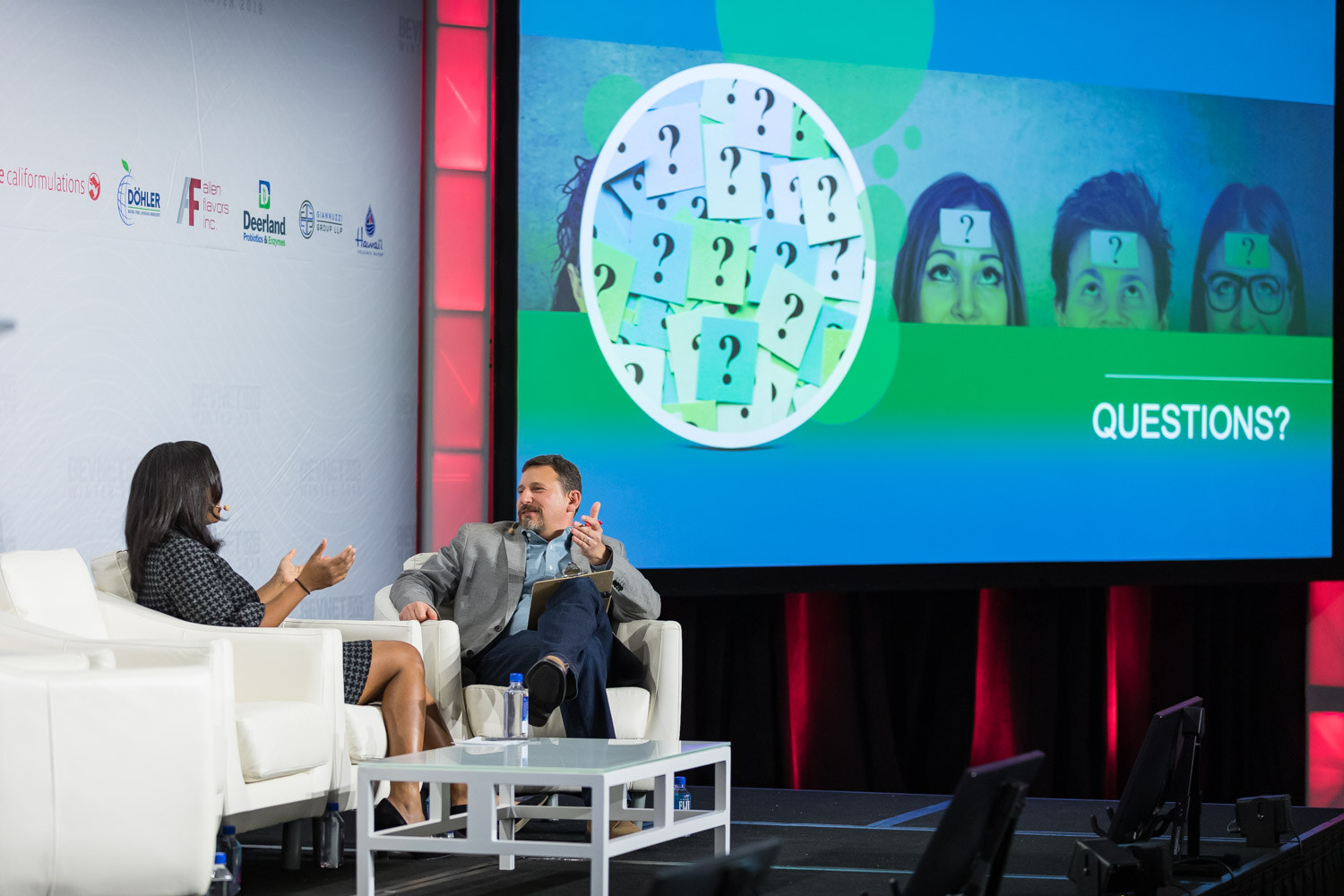Woman and man in armchairs on stage with a screen showing question marks and the word Questions.
