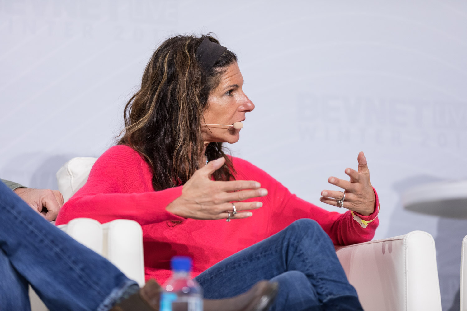 Woman in a red top gesturing while seated in a white armchair on stage.