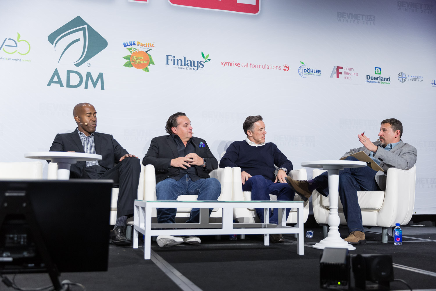 Four men seated in white chairs on a stage around a low coffee table.