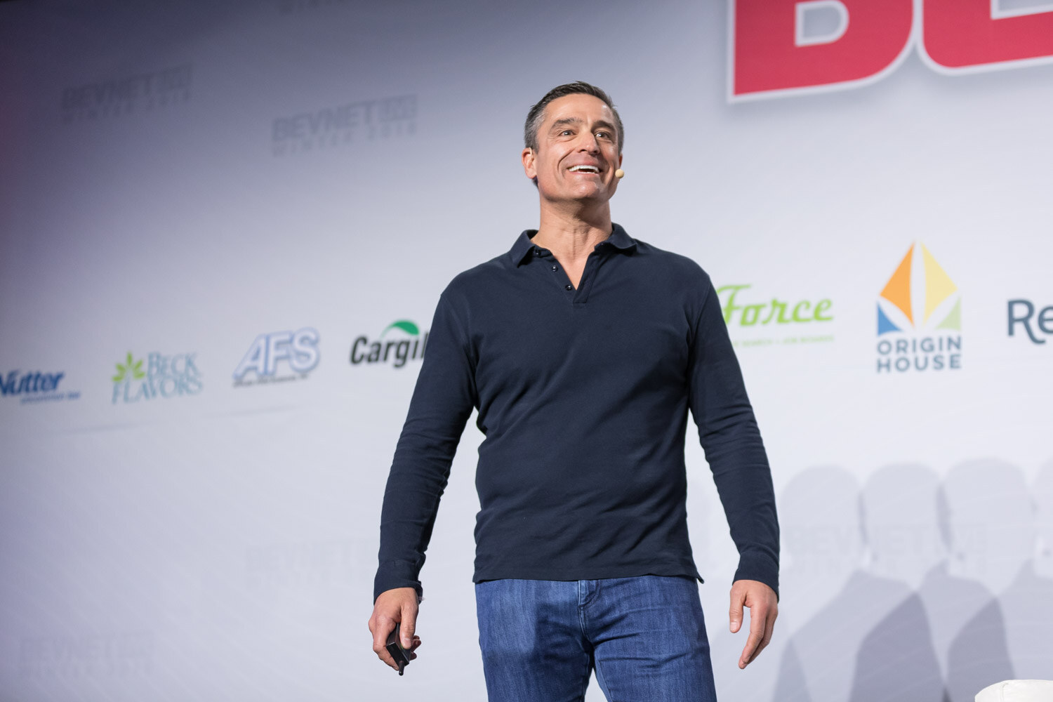 Man in a navy long-sleeve shirt holding a slide clicker with sponsor logos behind him.