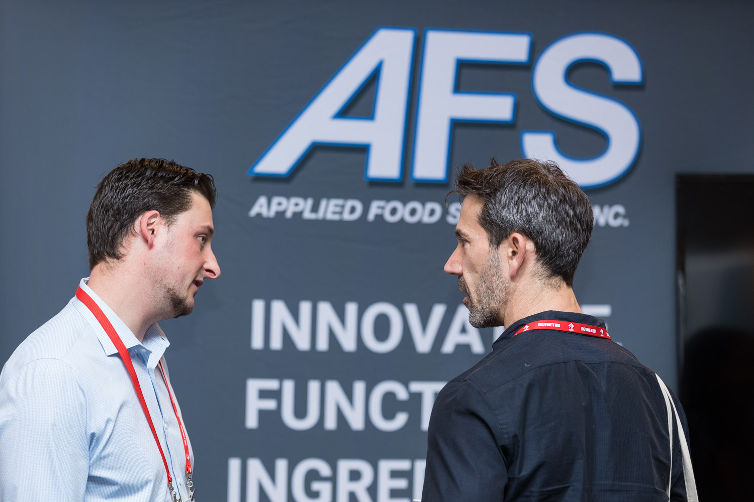 Two men with lanyards talking in front of a dark wall reading AFS Applied Food Sciences.