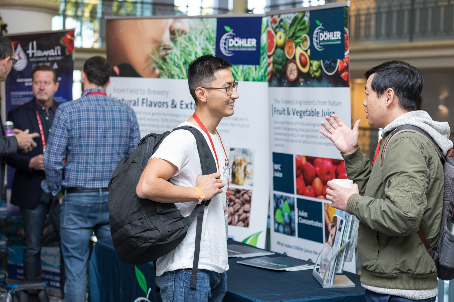 Man with a backpack talking to a man in an olive jacket in front of a Dohler banner.